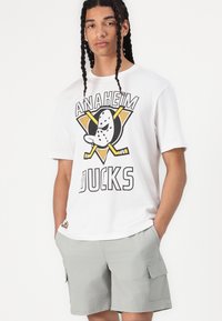 Fehér pamut póló az "Anaheim Ducks" logójával, amely egy rajzfilm figura. Szürke cargo sorttal párosítva. Rövid ujjú, laza szabású.