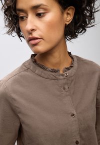 Camicia marrone, in tessuto strutturato, con colletto arricciato e un leggero luccichio, caratterizzata da un collo rotondo e con il primo bottone allacciato.