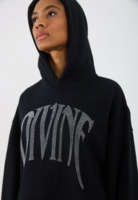 Hanorac negru cu un croi supradimensionat, având o grafică metalică cu strasuri "DIVINE" pe față. Material moale, cu glugă ajustabilă.