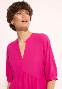 Fuksjowa sukienka z dekoltem w kształcie litery V i krótkimi bufkowymi rękawami, z wąską talią i małym guzikiem przy dekolcie, gładka tekstura materiału.