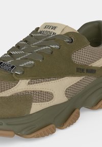 Gros plan d'une basket Steve Madden vert olive et beige avec panneaux en mesh, lacets à motifs et étiquette métallique de marque sur fond blanc.