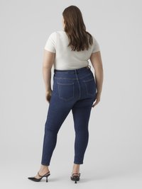 Donkerblauwe skinny jeans met hoge taille en een gladde textuur, voorzien van achterzakken en een strakke pasvorm, gecombineerd met een crème kleurig aangesloten t-shirt.
