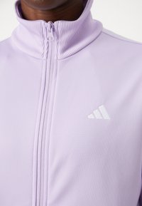 Ανοιχτό μωβ ζακέτα με φερμουάρ και ψηλό γιακά, φτιαγμένη από λείο ύφασμα. Διαθέτει λευκό λογότυπο Adidas στην αριστερή περιοχή του στήθους.