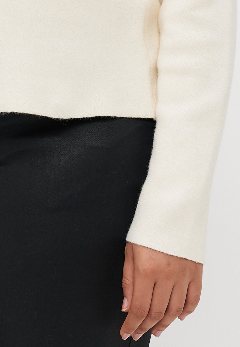 Cremfarbener Pullover mit weicher Textur, leicht verkürztem Schnitt und eng anliegenden langen Ärmeln, kombiniert mit einem schwarzen Kleidungsstück darunter.