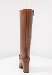 Bota de cuero marrón hasta la rodilla con un tacón grueso, que presenta una textura suave, costuras verticales y un acento decorativo en la parte trasera.