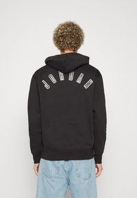 Felpa con cappuccio nera presenta la scritta "JORDAN" in lettere bianche arcuate sul retro, con polsini e orlo a costine, indossata con jeans azzurri chiari.
