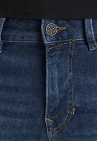 Krupni plan tamnoplavih traperica s smešnim šavom, metalnim dugmetom ugraviranim s "PME Legend Jeans" i petljama za pojas.
