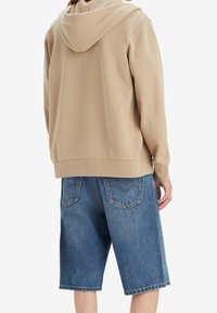 Beige huvtröja med en avslappnad passform, ihopkopplad med blå denim bermudashorts som har två bakfickor och en fransad fåll.