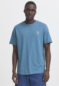 Blå bomulds T-shirt med rund halsudskæring og korte ærmer. Har et lille hvidt grafik på venstre bryst. Båret med denim shorts.