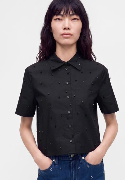 Jeune femme aux longs cheveux foncés portant une chemise noire à manches courtes boutonnée avec des ornements en perles et un jean bleu avec des détails en perles.