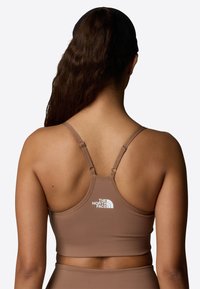 Sujetador deportivo marrón con una textura suave, tirantes ajustables y un logo blanco en la parte posterior. Diseño minimalista, adecuado para ropa deportiva.