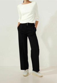 Frau in einem weißen Langarmoberteil, hoch taillierten, schwarzen, weiten Hosen mit Knöpfen und weißen Sneakern, die die Hände in den Taschen hat.