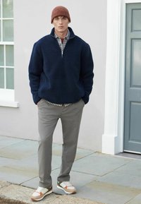 Marineblå fleece-pullover med halv lynlås, grå bukser og multicolore sneakers. Brun strikket beanie. Uformel udendørsindstilling.
