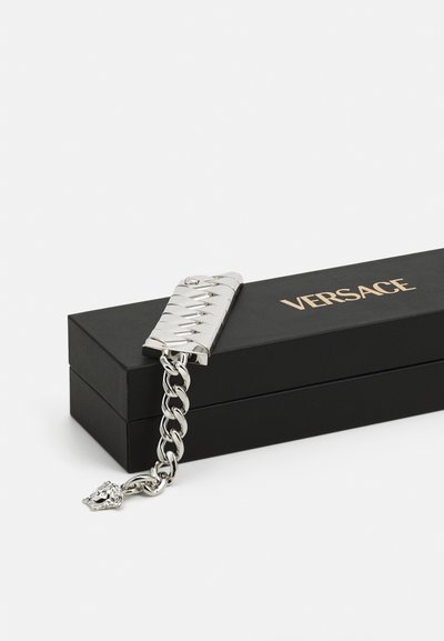 Ezüst fém karkötő szövet mintával, csattal és láncszemekkel, mellette egy fekete doboz, amelyen aranybetűkkel "VERSACE" felirat található.