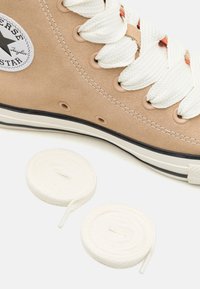 Sapatilhas de cano alto em suede bege com atacadores brancos, sola de borracha com detalhe em preto, e um emblema circular. Inclui dois pares adicionais de atacadores brancos.