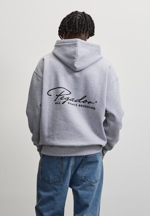 Persoon met gevlochten haar die een grijze hoodie met zwarte tekst "Pegador All Rights Reserved" draagt, gecombineerd met een blauwe spijkerbroek, met de rug naar de kijker.
