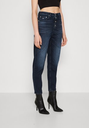 Vaqueros de mezclilla de color azul oscuro con cintura alta, diseño de cinco bolsillos y cierre de botón. Combinados con botas de tacón alto negras hasta el tobillo.