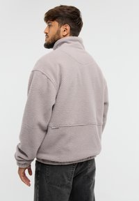 Felpa in pile di colore grigio chiaro, caratterizzata da un colletto alto, una vestibilità comoda e un tessuto testurizzato. La vista posteriore mostra l'orlo a costine e il design senza cuciture.