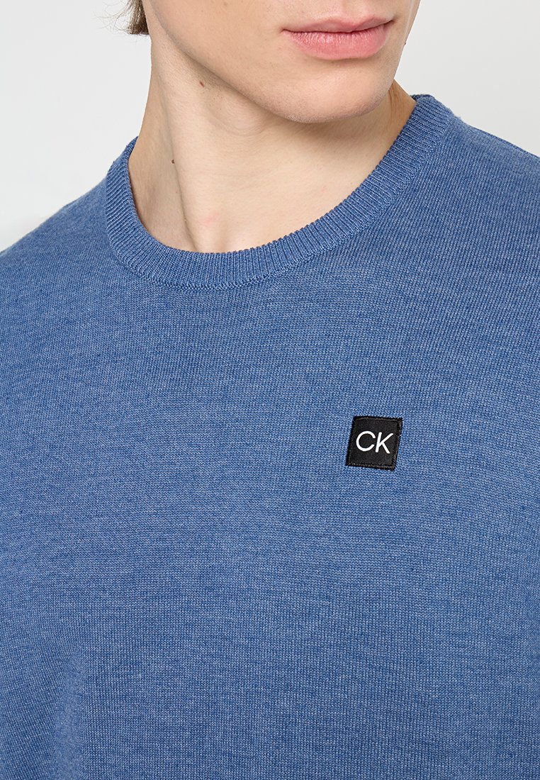 Un pull bleu à col rond en tissu doux, arborant une petite étiquette noire avec "CK" sur le côté supérieur gauche. Texture lisse et design simple.