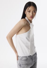 Top bianco senza maniche con righe verticali blu e un nodo al collo, abbinato a jeans grigi chiari con borchie decorative.