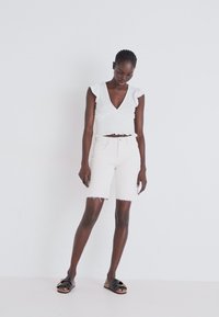 Haut blanc à manches courtes avec des bords volantés, associé à un short en denim blanc avec des ourlets effilochés. Sandales noires à enfiler avec des clous.