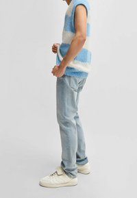 Pull sans manches à rayures bleues et blanches, fabriqué dans un matériau duveteux, associé à un jean bleu clair et des baskets blanches.