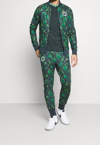 Modelo masculino con chándal de color verde oscuro con patrones de hojas en verde brillante y amarillo, zapatillas Nike blancas, de pie frente a un fondo claro.