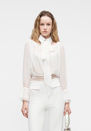 Kobieta w przezroczystej białej bluzce z kołnierzykiem z kokardą i białych spodniach z wysokim stanem, trzymająca małą białą torebkę na jednolitym tle.