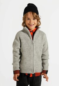 Szary prążkowany sweter na zamek z wysokim kołnierzem, noszony na pomarańczowo-czarnej koszuli w kratę. Dziecko ma na sobie czarną dzianinową czapkę.