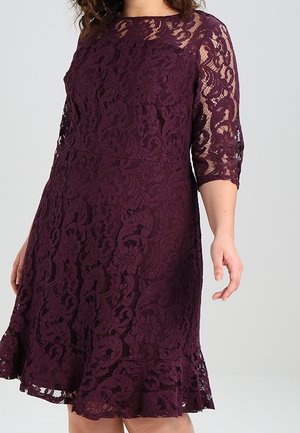 Kobieta w kremowej koronkowej sukience do kolan z rękawami do połowy przedramienia i rozkloszowanym dołem na szarym, jednolitym tle.