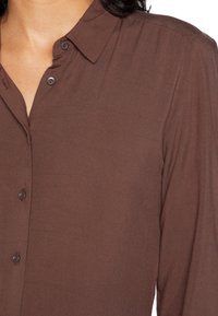 Camicia marrone con bottoni, realizzata in tessuto morbido, dotata di colletto, maniche lunghe e bottoni marroni scuri lungo la parte anteriore.