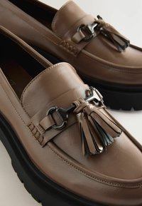 Bruna läderloafers med en kraftig svart sula, prydda med en dekorativ tofs och silverdetaljer framtill. Slät textur.
