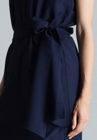 Femme portant une robe bleu marine avec une ceinture en tissu large nouée en un nœud à la taille, bras droit visible sur un fond gris uni.
