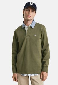 Long-sleeve olive green polo with a buttoned collar, layered over a light blue shirt, paired with beige pants. Logo accent on chest.
