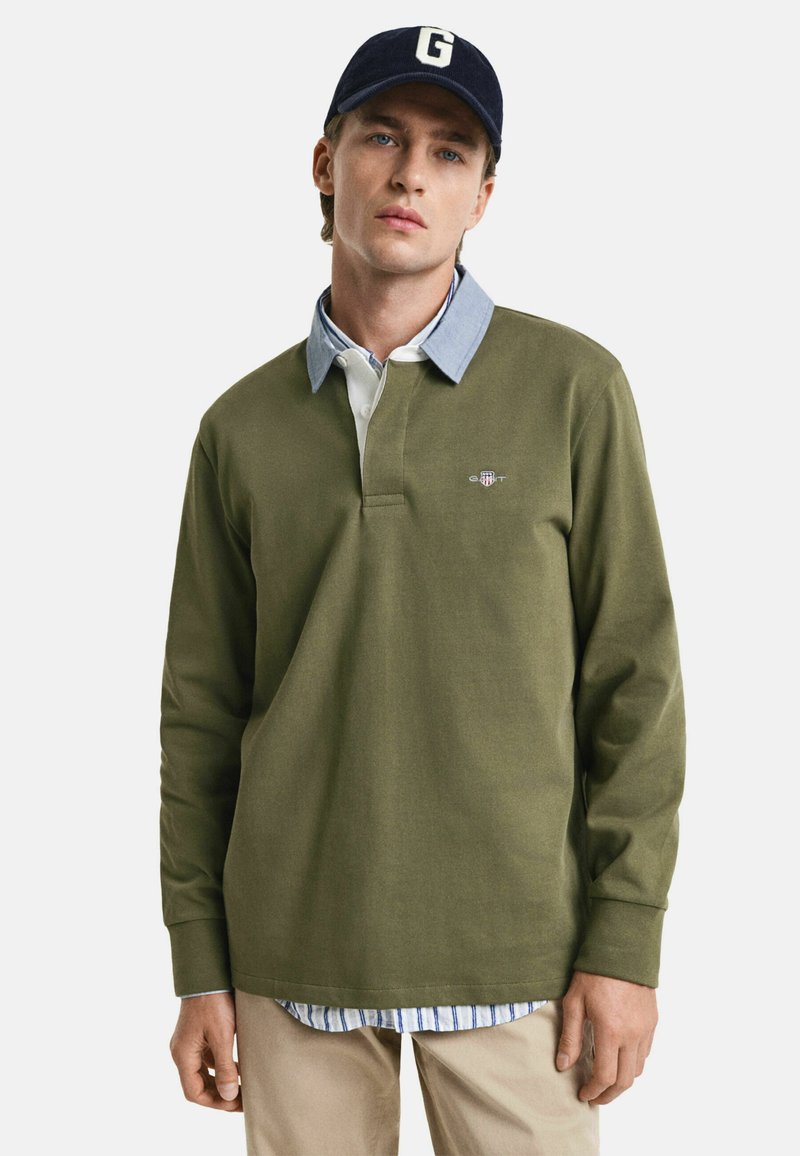 Long-sleeve olive green polo with a buttoned collar, layered over a light blue shirt, paired with beige pants. Logo accent on chest.
