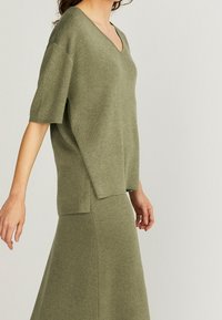 Olivgrön stickad topp med korta ärmar och V-ringning, tillsammans med en matchande kjol. Båda har en mjuk textur och en avslappnad passform.