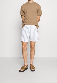 T-shirt marron à manches courtes associé à un short de bain rayé blanc avec un logo rouge. Porté avec des sandales à lanières marron.