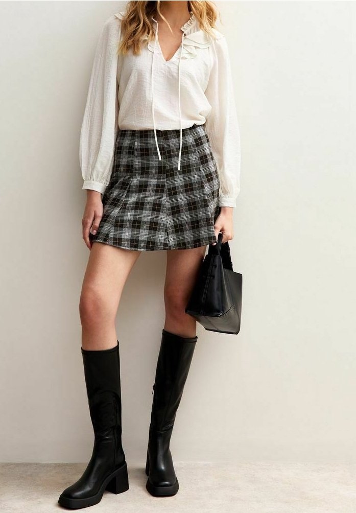 Chemisier blanc à col volanté, jupe écossaise grise et noire, bottes en cuir noires montant jusqu'aux genoux, tenant un petit sac à main noir.