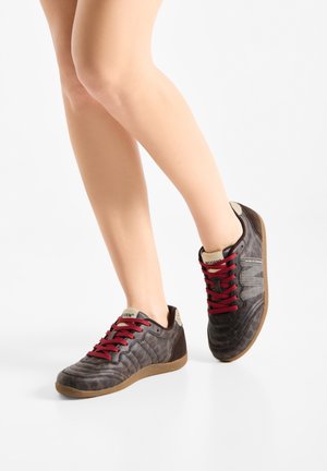 Gambe con sneakers marroni con lacci rossi e linguetta beige, in piedi su un pavimento bianco, un piede leggermente sollevato.