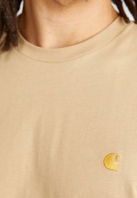 T-shirt in cotone beige con piccolo logo dorato ricamato sulla parte sinistra del petto. Maniche corte e scollo rotondo.