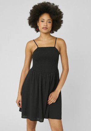 Jeune femme aux cheveux bouclés naturels portant une robe noire sans manches à fines bretelles, debout devant un fond clair uni.