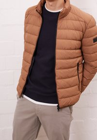 Giacca a vento marrone con texture trapuntata e colletto alto, abbinata a un maglione blu navy a collo rotondo e pantaloni beige chiaro. Tasche con zip visibili.