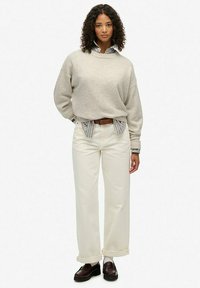 Beige trui met afgekapte schouders over een blauw gestreept overhemd, gecombineerd met witte broek met wijde pijpen en bruine loafers.