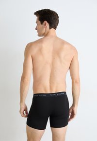 Boxers en coton noirs avec une large ceinture élastique arborant le logo "TOMMY HILFIGER" en blanc et rouge. Le modèle est montré de dos.