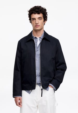 Mężczyzna z kręconymi włosami nosi rozpiętą czarną kurtkę na niebieskiej, w paski koszuli zapinanej na guziki i białych spodniach, stoi na jednolitym białym tle.