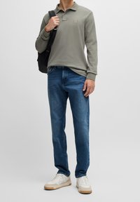 Ljust grå långärmad polo med en bröstlogotyp, i kombination med blå jeans och vita sneakers, hållande en svart ryggsäck.