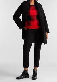 Personne portant un manteau noir texturé, un pull à carreaux rouge et noir, un pantalon noir retroussé et des bottes noires brillantes à lacets, debout.