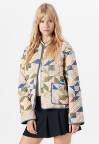 Giacca imbottita con un design a patchwork caratterizzato da motivi triangolari in blu, verde, rosa e crema. Bordo blu e dettaglio di tasca frontale.