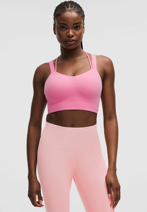 Vrouw met gevlochten haar, die een roze sportbeha en leggings met hoge taille draagt, staande tegen een effen lichte achtergrond.