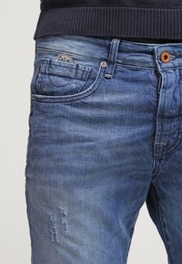 Jeans de denim azul con un lavado desgastado, un detalle rasgado en el muslo y un parche redondo con logo en el bolsillo trasero.